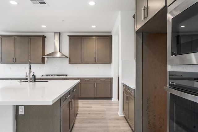 kitchen with a center island with sink, stainless steel appliances, wall chimney exhaust hood, light hardwood / wood-style flooring, and sink