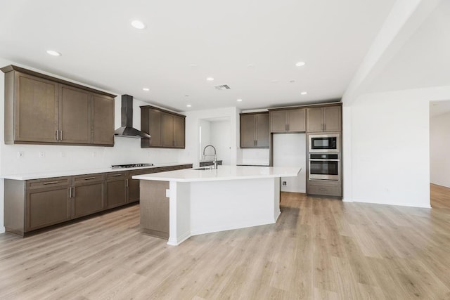 kitchen featuring wall chimney range hood, oven, gas cooktop, a kitchen island with sink, and built in microwave