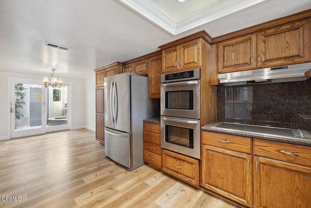 kitchen with an inviting chandelier, appliances with stainless steel finishes, under cabinet range hood, brown cabinets, and decorative backsplash