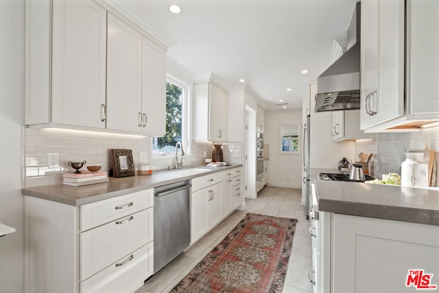 kitchen featuring stainless steel dishwasher, sink, wall chimney range hood, and white cabinetry