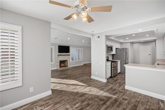 unfurnished living room with ceiling fan, dark wood-type flooring, a tray ceiling, and a stone fireplace