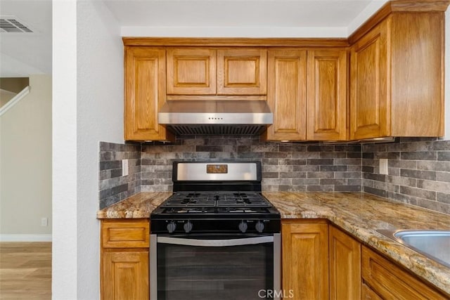 kitchen with light stone counters, tasteful backsplash, ventilation hood, stainless steel range with gas cooktop, and brown cabinetry