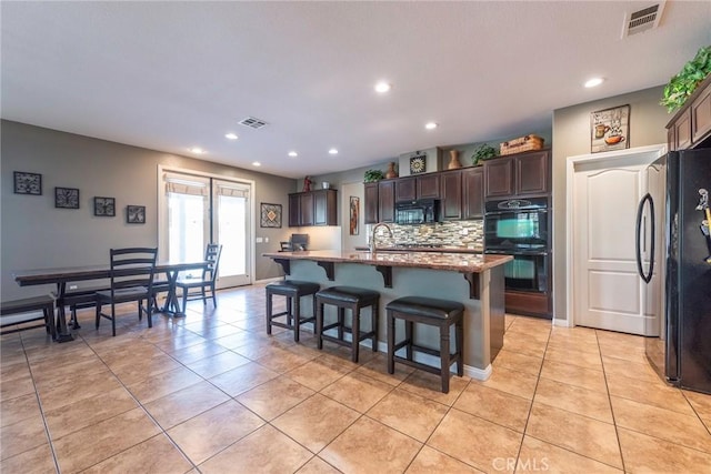 kitchen featuring light tile patterned floors, dark brown cabinets, a center island with sink, and black appliances