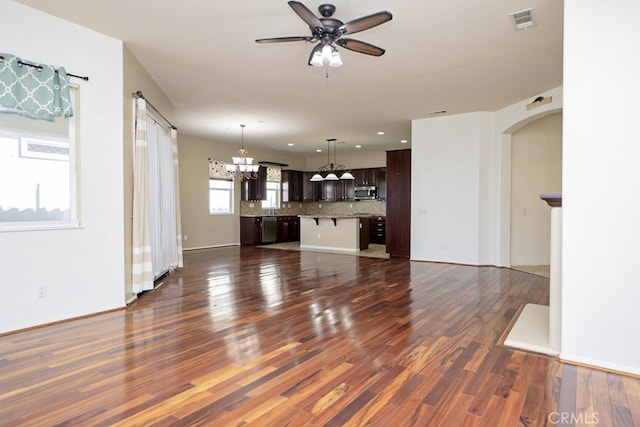 unfurnished living room with arched walkways, ceiling fan with notable chandelier, dark wood-type flooring, visible vents, and baseboards