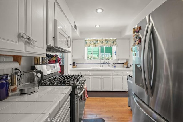 kitchen with appliances with stainless steel finishes, sink, white cabinets, tile countertops, and light hardwood / wood-style floors