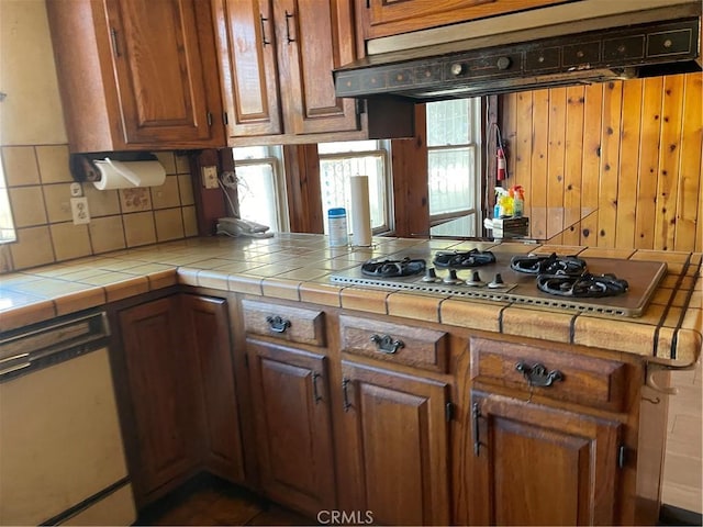 kitchen with tile countertops, stainless steel gas cooktop, a peninsula, dishwasher, and brown cabinetry