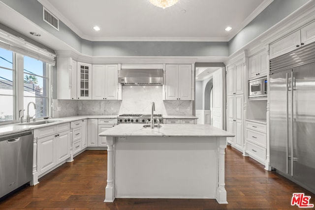 kitchen with wall chimney range hood, sink, built in appliances, an island with sink, and white cabinets