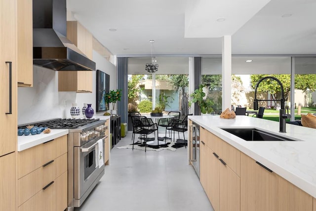 kitchen with wall chimney range hood, light brown cabinets, range with two ovens, sink, and light stone counters