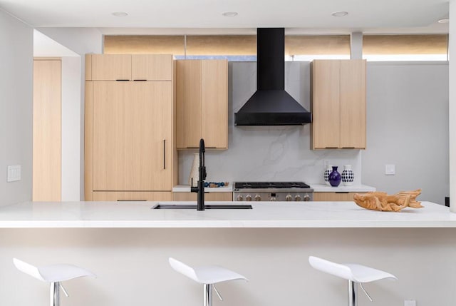 kitchen featuring tasteful backsplash, a kitchen bar, wall chimney range hood, and light brown cabinets