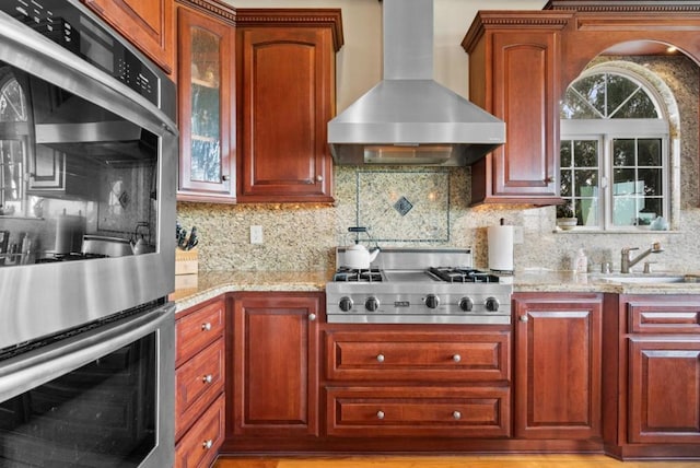 kitchen featuring light stone counters, appliances with stainless steel finishes, wall chimney exhaust hood, and sink