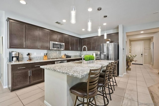 kitchen featuring light stone countertops, dark brown cabinetry, appliances with stainless steel finishes, decorative light fixtures, and a kitchen island with sink