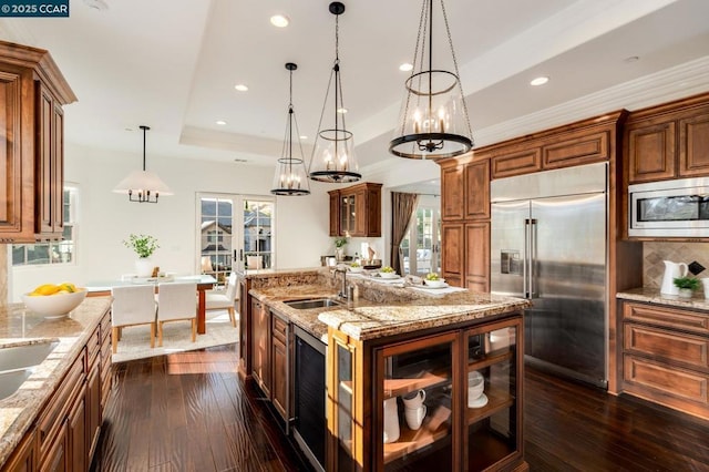 kitchen featuring decorative light fixtures, built in appliances, an island with sink, sink, and a tray ceiling