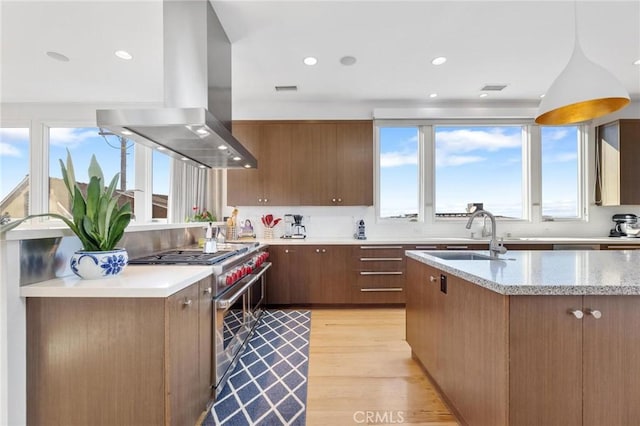 kitchen with high end stove, a sink, brown cabinetry, island exhaust hood, and pendant lighting