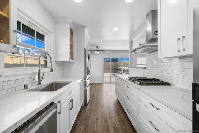 kitchen with light stone countertops, stainless steel appliances, wall chimney range hood, white cabinetry, and sink