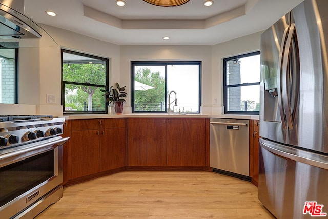 kitchen with appliances with stainless steel finishes, a tray ceiling, extractor fan, and light hardwood / wood-style flooring