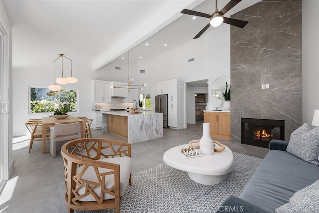 living room featuring beam ceiling, a high end fireplace, ceiling fan with notable chandelier, and a towering ceiling