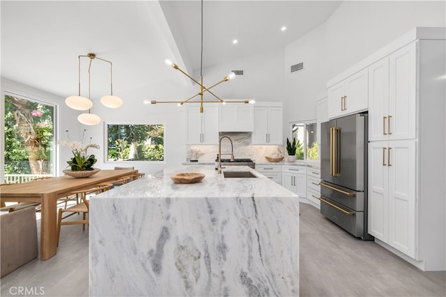 kitchen with high quality fridge, white cabinets, hanging light fixtures, a kitchen island with sink, and light stone counters