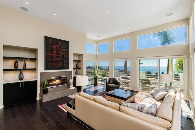 living room featuring a towering ceiling, built in shelves, dark hardwood / wood-style flooring, a fireplace, and a water view