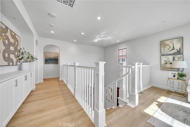 hallway featuring arched walkways, light wood finished floors, recessed lighting, visible vents, and an upstairs landing