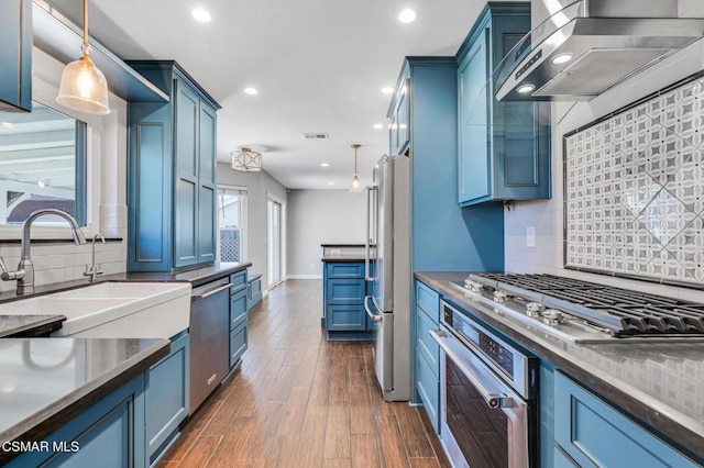 kitchen with appliances with stainless steel finishes, wall chimney exhaust hood, blue cabinets, and pendant lighting