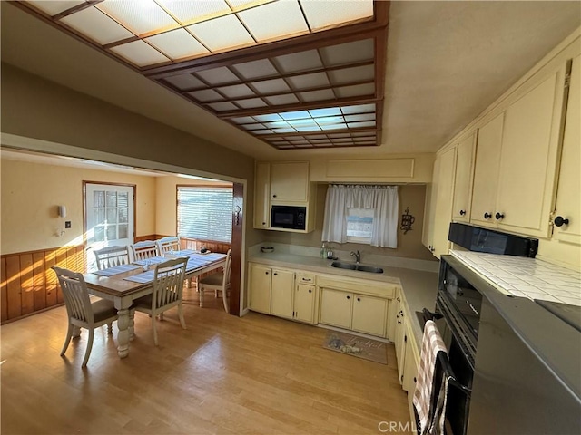 kitchen with black microwave, tile counters, sink, wood walls, and light hardwood / wood-style floors