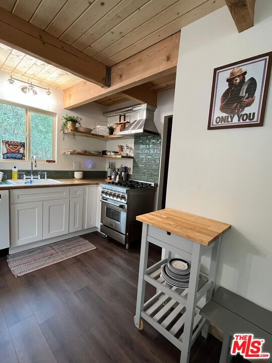 kitchen with white cabinets, wall chimney range hood, sink, stainless steel stove, and beam ceiling