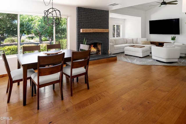 dining space featuring a brick fireplace, wood-type flooring, ceiling fan with notable chandelier, and plenty of natural light