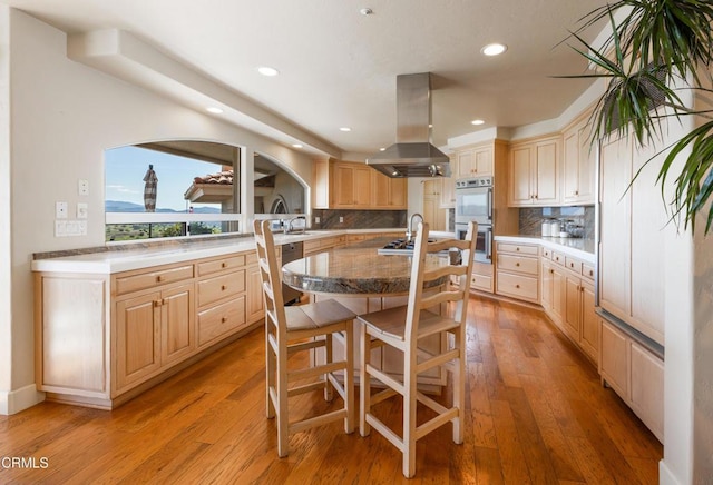 kitchen with light brown cabinetry, island range hood, backsplash, and double wall oven