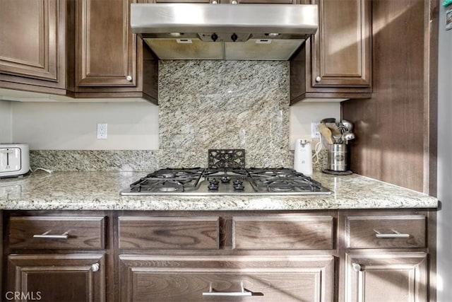 kitchen with backsplash, exhaust hood, stainless steel gas stovetop, dark brown cabinetry, and light stone counters