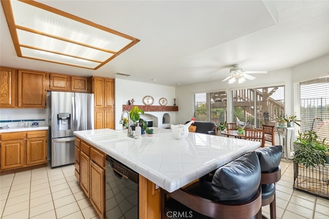 kitchen with tile countertops, black dishwasher, stainless steel fridge, light tile patterned floors, and a center island with sink