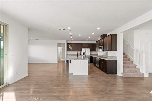 kitchen with appliances with stainless steel finishes, hardwood / wood-style flooring, light stone counters, and a kitchen island with sink