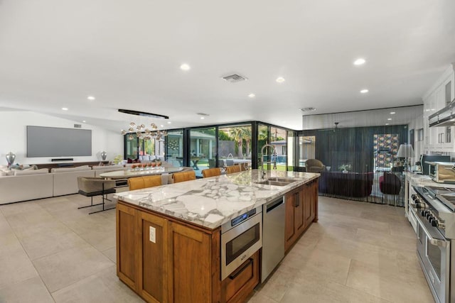 kitchen with stainless steel appliances, a kitchen island with sink, light stone counters, expansive windows, and sink