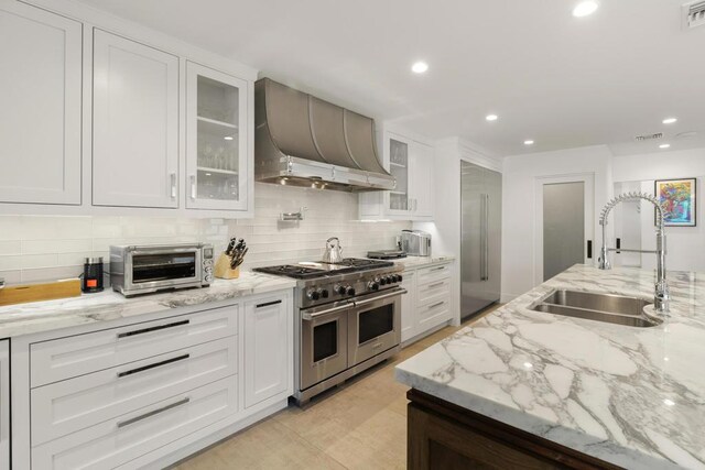 kitchen with wall chimney range hood, range with two ovens, light stone countertops, white cabinets, and sink