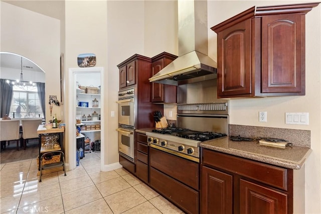 kitchen with wall chimney range hood, light tile patterned flooring, a high ceiling, appliances with stainless steel finishes, and a chandelier