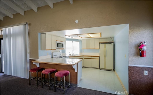 kitchen with kitchen peninsula, sink, white appliances, a breakfast bar, and beamed ceiling