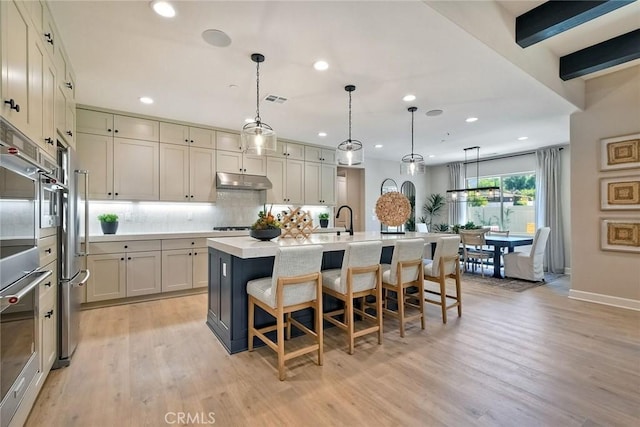 kitchen with beamed ceiling, light hardwood / wood-style flooring, a kitchen island with sink, and hanging light fixtures