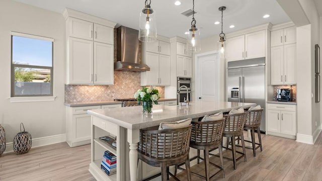 kitchen featuring appliances with stainless steel finishes, hanging light fixtures, an island with sink, white cabinets, and wall chimney exhaust hood