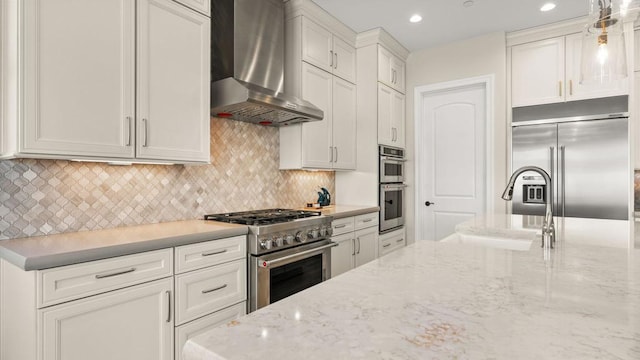 kitchen featuring hanging light fixtures, high quality appliances, wall chimney range hood, light stone countertops, and white cabinets