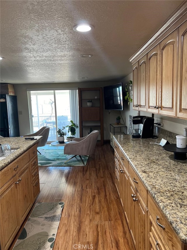 kitchen with brown cabinetry, dark wood finished floors, light stone counters, freestanding refrigerator, and a textured ceiling