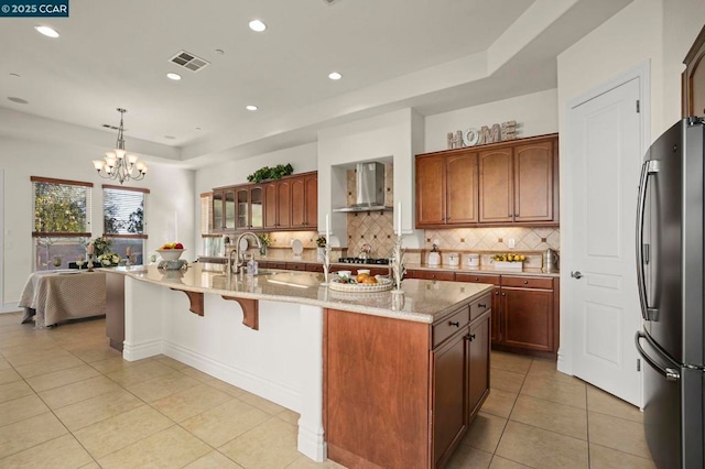 kitchen with a center island with sink, hanging light fixtures, wall chimney exhaust hood, stainless steel fridge, and a chandelier