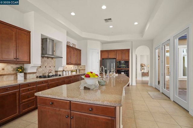 kitchen featuring tasteful backsplash, wall chimney range hood, a kitchen island with sink, stainless steel fridge, and light stone counters