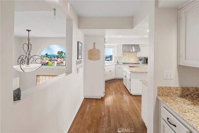 kitchen with wall chimney range hood, tasteful backsplash, light stone countertops, white cabinets, and decorative light fixtures