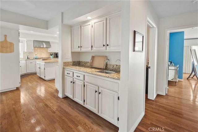 kitchen with white cabinetry, sink, light stone counters, wall chimney range hood, and light hardwood / wood-style flooring