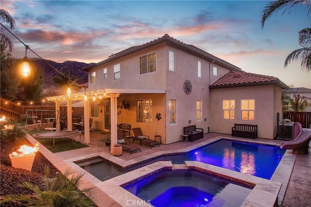 back house at dusk featuring a patio area, a pool with hot tub, a trampoline, and a pergola