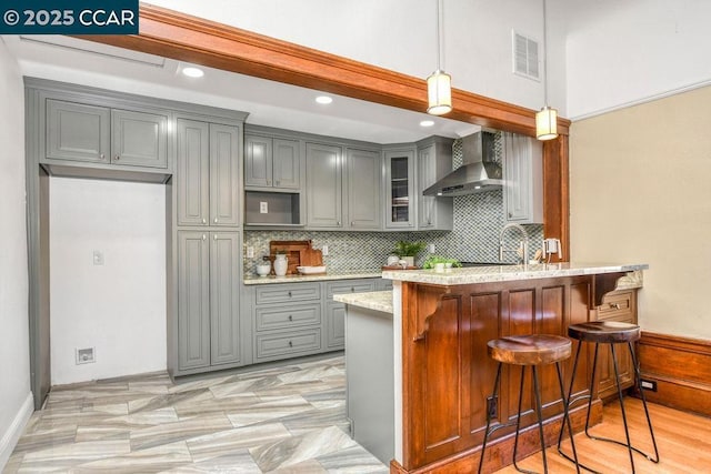 kitchen with pendant lighting, wall chimney exhaust hood, kitchen peninsula, gray cabinetry, and a breakfast bar area