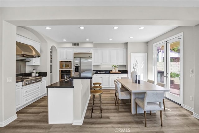 kitchen featuring white cabinets, a center island with sink, appliances with stainless steel finishes, and wall chimney exhaust hood