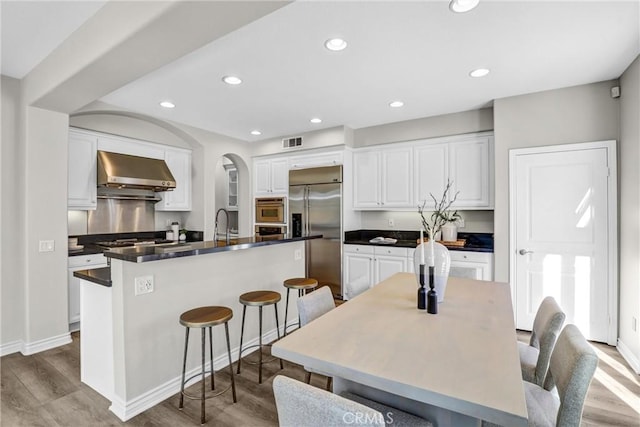 kitchen featuring stainless steel appliances, a center island with sink, wall chimney range hood, light wood-type flooring, and white cabinets