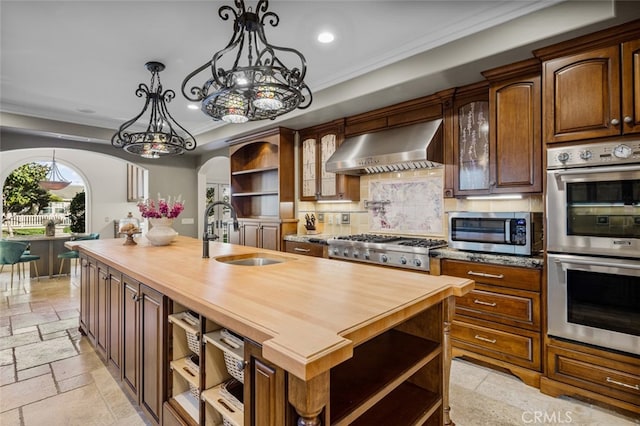 kitchen with wall chimney range hood, a center island with sink, sink, appliances with stainless steel finishes, and wood counters