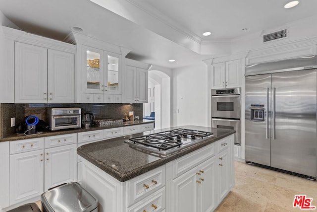 kitchen with appliances with stainless steel finishes, backsplash, dark stone countertops, ornamental molding, and white cabinets