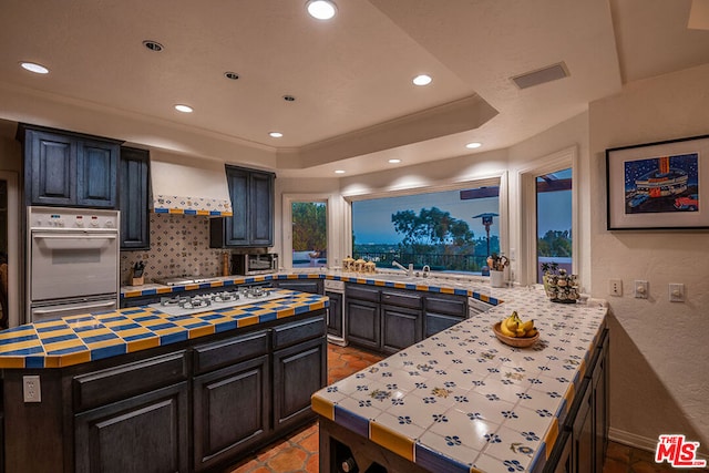 kitchen featuring appliances with stainless steel finishes, custom exhaust hood, kitchen peninsula, a raised ceiling, and light tile patterned floors